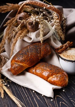 Baked sweet rolls sprinkled with poppy seeds lie on burlap on country table. Two rolls and old enameled cup with coffee and cinnamon near wicker basket with poppies and natural dried herbs.