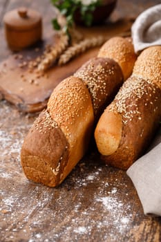 Close up of multigrain buns on wooden table in rustic style. Bread products with sesame seeds on blurred rustic background with spikelets. Pastries and bread in bakery.