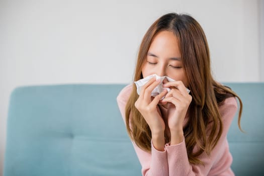 Ill Asian young woman cold covered with blanket sitting on sofa and sneeze with tissue paper at home, Female sick allergic blowing nose sneezing in tissue at home, Flu health care