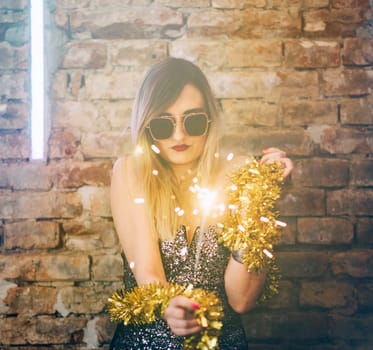 Young sexy woman celebrating event party with fireworks and confetti in night disco club. Beautiful woman with retro sunglasses in front of brick wall and disco lights.