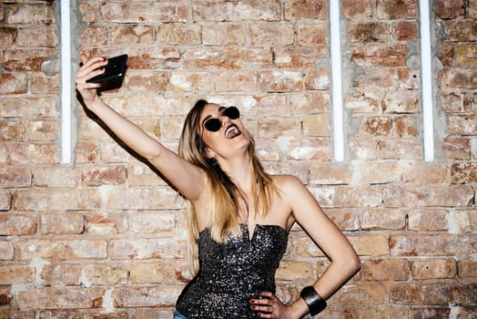 Young sexy woman posing in nightclub and taking selfie with her smartphone. Glamour stylish beautiful girl having fun in front of disco brick wall