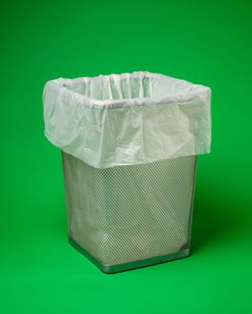 Empty metal trash can with white environmentally friendly biodegradable garbage bag stands on green background. Environmental safety concept. Close up. Place for text.