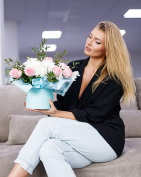 Stylish young blonde woman sitting on sofa with beautiful bouquet of flowers in her hands. Beautiful elegant woman looking at gift box filled with fresh white and pink flowers. Flower shop advertising