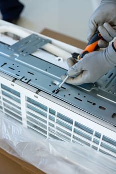 Male installer or repairman is going to install air conditioner unit. Close up of hands of young skilled male worker in household gloves who fastens bolts on air conditioner with screwdriver.