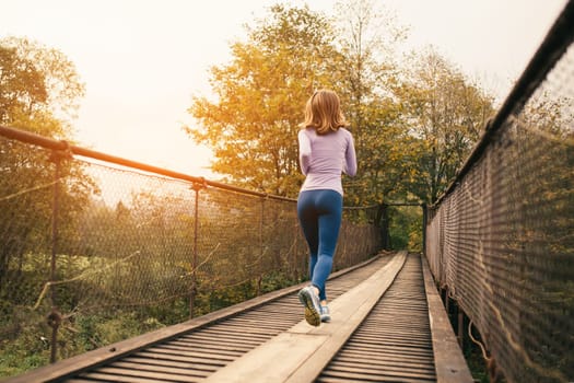 Fitness and sports motivation. Rear view of a sporty woman runner running over an old suspension bridge on a cool autumn day. Outdoor cardio exercise concept.