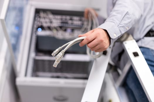 Server hardware setup. Male worker configures local network by holding cables and plugs to connect to server. Close up of plug in hand of man standing on ladder.