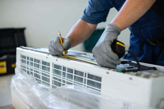 Male technician makes mark with pencil when measuring air conditioner unit. Close up of man's hand in protective gloves holds pencil and puts marks measuring air conditioner with tape measure.