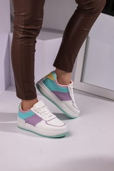 Cropped image of new collection of fashionable bright sports shoes for teens. White laceup sneakers with turquoise and purple inserts on legs of young woman dressed in brown pants.