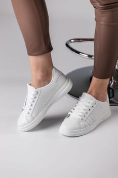 Close up of fashionable white sneakers on legs of girl on white background in studio. Young woman in brown pants demonstrates new collection of sports shoes with laces and with breathable holes.