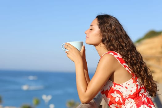 Side view portrait of a woman in red dress relaxing smelling coffee on the beach