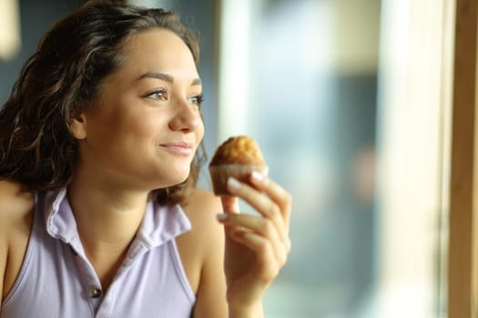 Happy woman holding a muffin in a restaurant and looking through a window