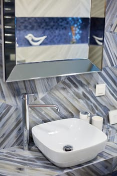 Modern washbasin with chrome faucet in stylish and bright bathroom of new house. Mirror hangs over sink in room with trendy tiles on walls. Concept of details.