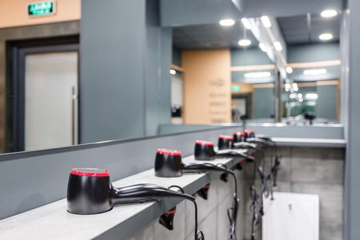 Close up of stand in washroom in gym on which are placed in row many hair dryers. Room with mirrors and hairdryers in gym or pool. Concept of services in gym or pool. Selective focus.