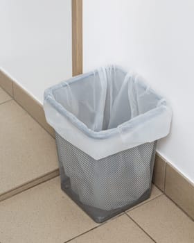 Close up of small trash can with recyclable garbage bag. Empty metal gray trash can with white bag inside stands on floor under wall indoors. Ecology concept.