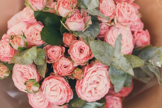 Pink spray roses with sprigs of green eucalyptus stacked together in one festive bouquet. Close up of bouquet of flowers wrapped in brown kraft paper. Concept of flower delivery for any holiday.