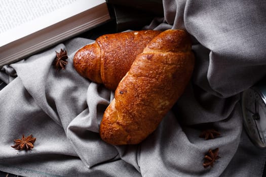 Close up of fresh pastries on gray burlap cloth near open book to read. Top view of two brown sweet baked buns lying next to books, retro clock and star anise. Vintage style view.