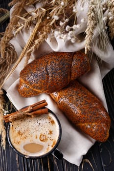 Fresh baked rolls with poppy seeds and coffee on rustic dark wooden table. Top view of two fresh buns on light burlap and enameled cup with coffee and cinnamon sticks near rye and wheat ears.