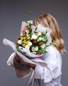 Woman in a delicate white shirt holds a bouquet of flowers with roses, tulips and greenery. Unknown blonde woman stands sideways on a gray background and holds flowers that cover her face.