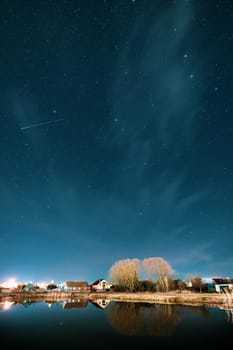 Belarus, Eastern Europe. Night Sky Stars Above Countryside Landscape With Lake Coast And Small Town Or Village. Natural Starry Sky Above Pond And Houses In Early Spring Night.