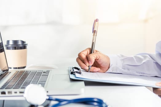 Closeup of doctor or nurse woman in uniform with stethoscope writing information of patient prescription in paperwork on clipboard and typing laptop computer for history record medical document report