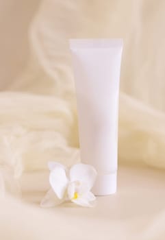 White Cream tube near orchid flowers and tulle on light beige close up. Packaging Mockup. Exotic natural cosmetics, romantic composition, everyday skincare routine..