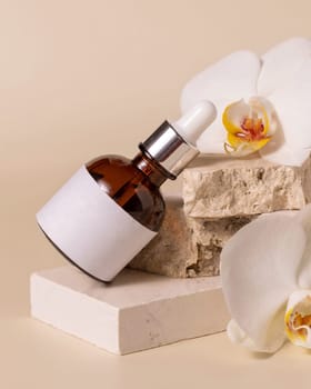 Brown glass dropper bottle with blank label laying on a stone near white orchid flowers on light beige, close up, mockup. Skincare beauty product, serum or essence. Exotic natural cosmetics..