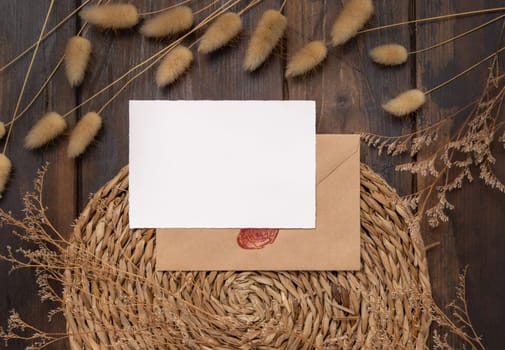 Blank card and envelope on wattled placemat on wood near Hare's Tail grass top view. Boho scene with horizontal paper card. Bohemian or close to nature greeting or Invitation mockup, place for text.
