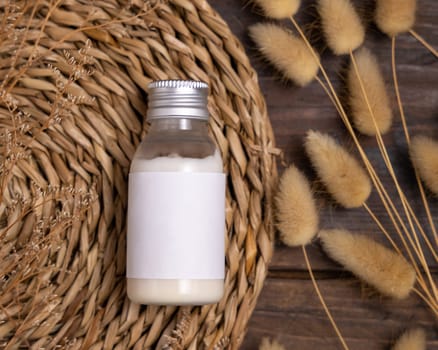 Cosmetic bottle on boho wattled placemat near Hare's Tail grass top view on wood, label mockup. Natural skincare product, cream or lotion. Bohemian eco friendly beauty flat lay.