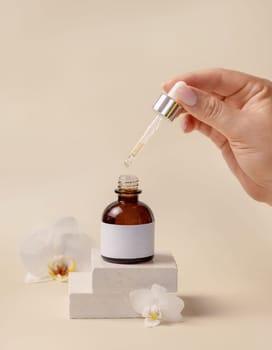 Hand holding pipette over opened cosmetic dropper bottle near white orchid flowers and stones on light beige, close up, mockup. Natural skincare product, serum or essential oil.