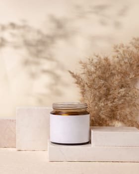 Glass cosmetic jar with blank label on stones near pampas grass on light beige close up, packaging mockup. Skincare beauty product, lotion or cleanser. Natural eco friendly cosmetics
