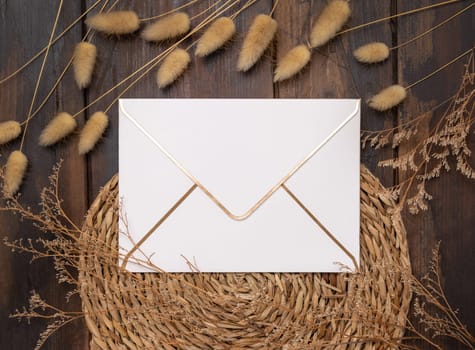 Blank envelope on wattled placemat on wood near Hare's Tail grass top view. Boho scene with horizontal paper envelope. Bohemian or close to nature greeting or Invitation mockup, place for text.