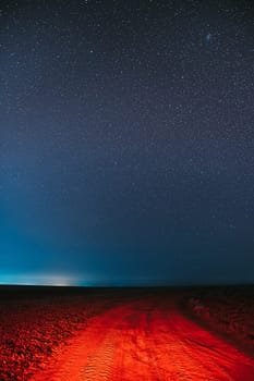 Night Starry Sky With Glowing Stars Above Country Road Is Lit In Red. Countryside Field Landscape.