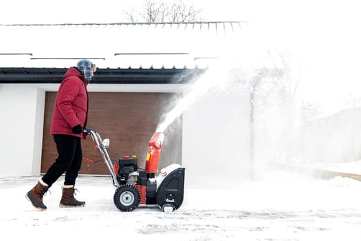 Snow blower powered by gasoline in action. Man outdoor in front of house using snowblower machine. Snow removal, thrower assistant in winter outside home. Young worker guy blowing snow during blizzard