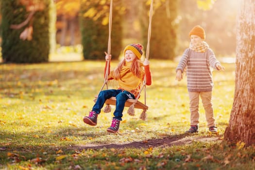 Couple of children playing in the autumnal park. Boy swinging girl on the swing. Concept of friendship and romantic relationship.