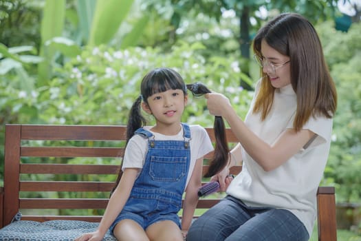 Asian smiling child schoolgirl with long dark hair and her mom in the morning, Happy family beautiful mother woman brushing her daughter hair while sitting outdoor at home, hairstyle care concept