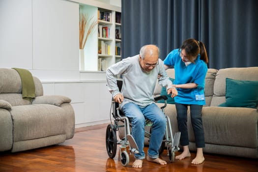 Asian nurse assisting helping senior man patient get up from wheelchair for practice walking at home, Smiling doctor support old man to getting up, help handicapped elderly stand up, healthcare