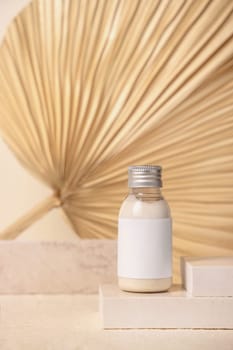 Small bottle with blank label on travertine stone against dry beige palm leaf close up, mockup. Skincare beauty product - cream or lotion. Natural organic cosmetics. Minimal eco friendly composition..