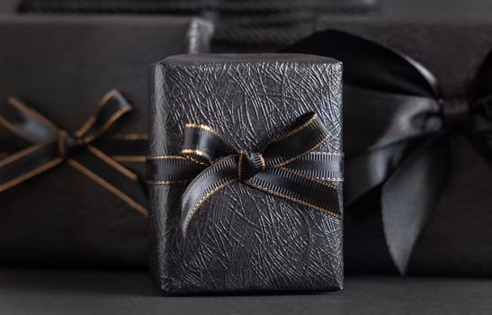 Black Gift Boxes with a ribbon bow on black close up. Wrapped presents for Dark themed Party, Christmas, Birthday, Anniversary. Black Friday concept. Monochrome composition