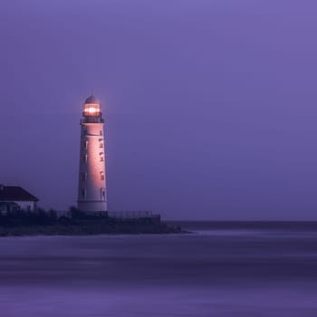 A lighthouse on the coast of the sea at dusk, its bright lamp is a landmark for ships. A lighthouse in the lilac twilight. Seascape, nature background.