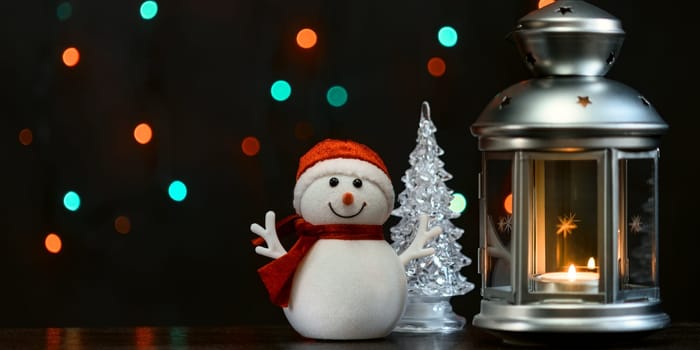 Christmas composition: a snowman, a lantern with candles and blurred bokeh lights in the background.