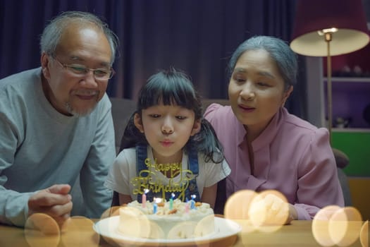 Happy Asian family grandparents and mother celebrating enjoy birthday party with cake of granddaughter Blowing candle at home in night time, Birthday surprise concept