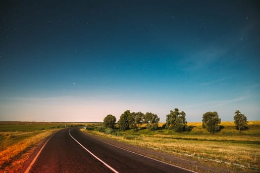 Blue Night Starry Sky Above Country Asphalt Road In Countryside And Green Meadow. Night View Of Natural Glowing Stars.