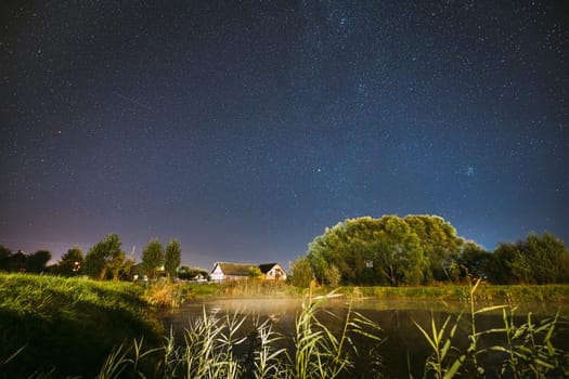 Evaporation Over River Lake Near Houses In Village. Milky Way Galaxy In Night Starry Sky Above Lake River Landscape At Night. Glowing Stars Above Summer Nature