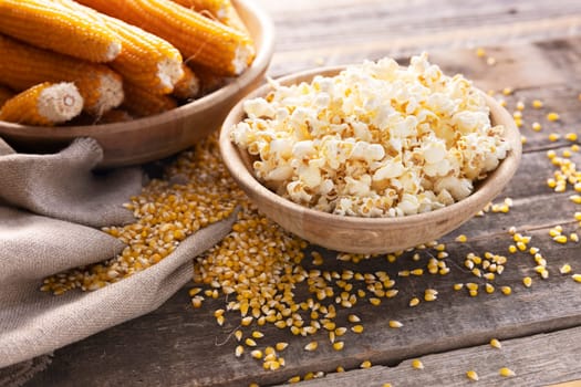 Wodden bowl of popcorn surrounded by popcorn kernels and popcorn on the cob