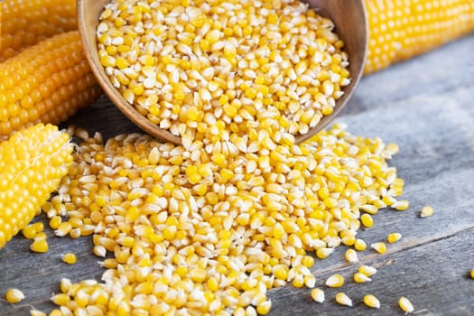Popcorn kernels spilling from wooden bowl onto talble with cobs of popcorn in background.