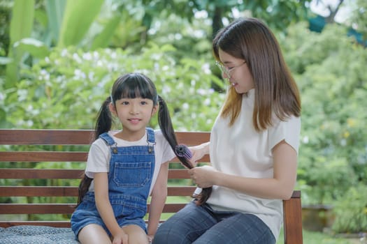 Happy family beautiful mother woman brushing her daughter hair while sitting outdoor at home, Asian smiling child schoolgirl with long dark hair and her mom in the morning, hairstyle care concept