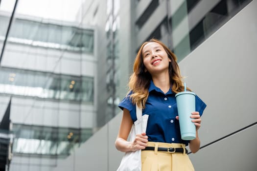 Asian beautiful business woman confident smiling with cloth bag holding steel thermos tumbler mug water glass she walking outdoors on street near modern building office, Happy female looking side away