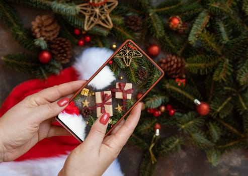 Woman's hand taking a mobile photo of christmas composition with Santa Claus hat, fir branches and holiday decorations top view. Blogger preparing content for social media using cell phone.