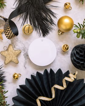 Paper round gift tag between fir tree branches and black and golden Christmas decorations on a white marble table top view. Mockup, copy space. Winter holiday flat lay with blank label