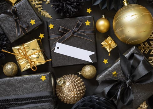 Black and golden Christmas Gift Boxes with a bow and ornaments close up, horizontal paper gift tag mockup, copy space. Dark winter composition with blank label card for Christmas, New Year, Birthday, Anniversary